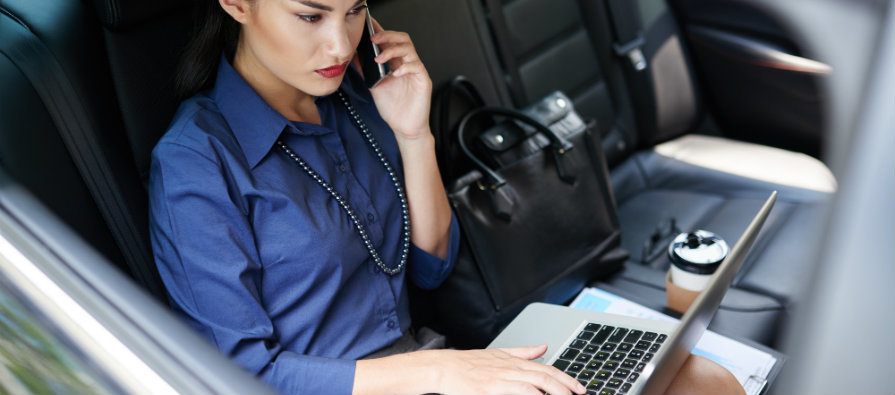 Mulher vestida como executiva utiliza notebook e celular no banco de trás do carro