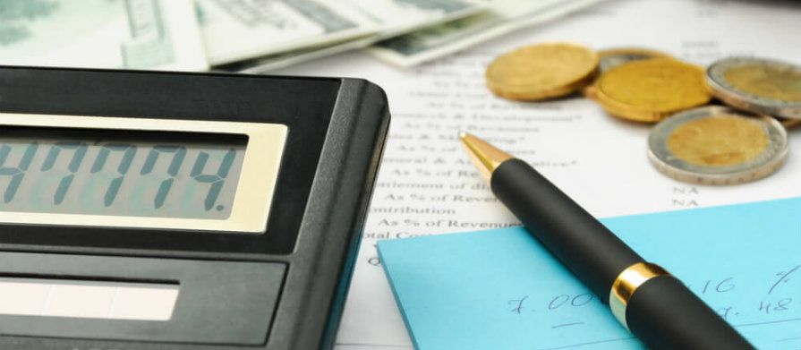 Papeis com contas, uma caneta, moedas empilhadas e uma calculadora ligada mostrando números no visor estão sobre a mesa.