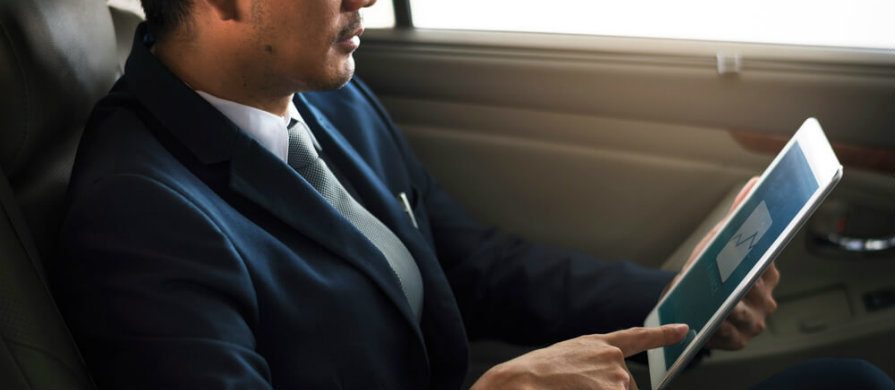 Homem analisa gráfico pelo seu tablet. Ele está sentado no banco traseiro de um carro e vestido de terno e gravata.