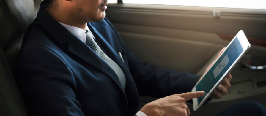 Homem analisa gráfico pelo seu tablet. Ele está sentado no banco traseiro de um carro e vestido de terno e gravata.