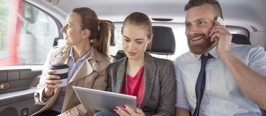 Três empresários. Duas mulheres e um homem estão sentados no banco traseiro de um carro. O homem fala ao celular, a mulher ao meio usa um tablet e a terceira olha pela janela do veículo que está em movimento.