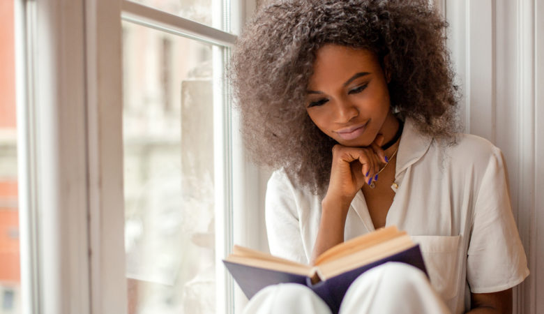Mulher sentada ao lado de uma janela lendo livros de investimentos.